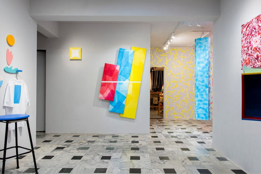 Installation view of a contemporary art exhibition featuring colorful geometric paintings and wall-based objects arranged across a gallery space, exploring CMY color relationships.