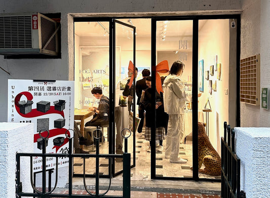 Exterior view of a small gallery during the Unboxing group exhibition, with the glass door open, visitors inside interacting with artworks, and an exhibition poster displayed at the entrance.